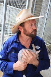 a photo of Kev our CEO. He has a rugged beard, wearing an akubra hat. Looking to the side. He is wering a blue SaVe the Bilby Fund button-up shirt and he is holding a bilby.