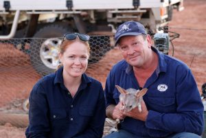 Fair-skinned Cass on the left with her red hair pulled back and black sunglasses on her head. She is smiling. Sitting next to her is Kev wear a cap and holding a bilby. Behind them is a ute on red outback soil. They both have dark blue button up shirts on to protect them from the outback sun.