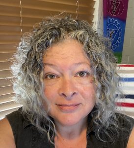 A selfie of Sara with her beautiful naturally white and grey, curly hair. She has kind eyes and a soft smile.